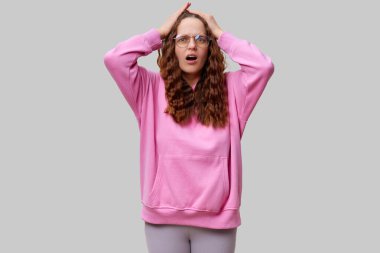 Kıvırcık saçlı ve gözlüklü bir kadın tarafsız bir ortamda duruyor, elleri başının üstünde şaşkın şaşkın bakıyor. Pembe bir kapüşonlu giyiyor ve beklenmedik haberlere tepki veriyor gibi görünüyor..