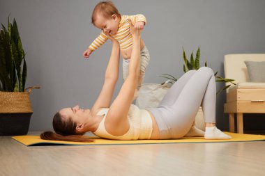 Parlak bir oturma odasında, bir anne yoga minderinde yatarken bebeğini kaldırarak fiziksel aktiviteye girişir. Bitkiler ve doğal ışık sıcak ve davetkar bir ortam yaratır..