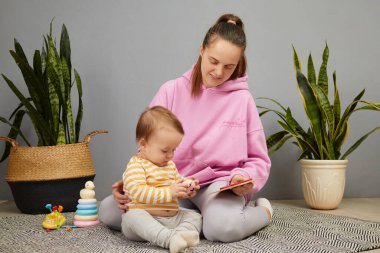 Bir anne, meraklı çocuğuna kitap okurken yumuşak bir halıya oturur. Kapalı alandaki bitkiler bu bağlanma anının tadını çıkarırken sıcak atmosfere tazelik katıyor..