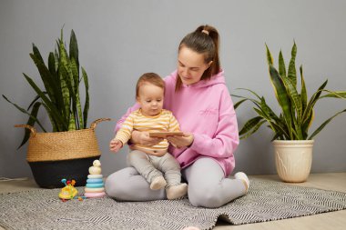 Meraklı bebeği yanında oynarken, bir anne elinde tabletle dokulu bir mindere oturur. Etrafları saksılarla çevrili, sıcak ve davetkar bir atmosfer yaratıyorlar..