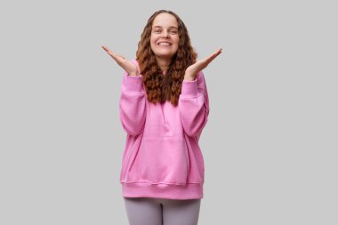 Kıvırcık saçlı neşeli bir kadın davetkar bir gülümsemeyle ellerini kaldırıyor. Pembe bir kapüşonlu giyiyor ve gri bir arka planın önünde pozitiflik yayıyor..