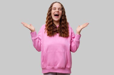 Dalgalı saçlı genç bir kadın pembe bir kapüşon giyerken heyecanla kollarını kaldırıyor. Yüzü sade bir arkaplan karşısında mutluluk saçıyor..