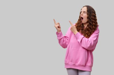 Açık gri bir ortamda pembe kapüşonlu genç bir kadın duruyor. Parmaklarını yana doğrultup heyecan ve enerji iletirken şaşırmış ve canlanmış görünüyor..