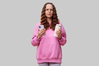 Pembe kapüşonlu bir kadın bir elinde akıllı telefon, diğer elinde kahve fincanı tutarak içeride duruyor. Kafası karışmış ya da meraklı görünüyor, kamera dışında bir şeye bakıyor..