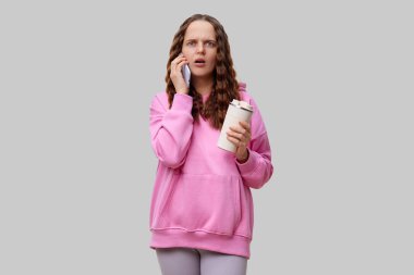 Elinde kahve fincanı tutarken telefonla konuşan pembe kapüşonlu bir kadın şok olmuş bir ifadeyle ayakta duruyor. Kıvırcık saçları o anın duygusal etkisini arttırıyor..