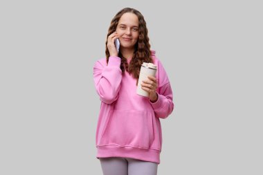 Genç bir kadın açık pembe bir kapüşonlu ve elinde kağıt bir kahve fincanı olan telefonunu kullanırken gülümsüyor. Tarafsız bir arkaplanda rahat ve neşeli görünüyor..