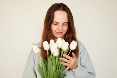 Bir kadın, yumuşak, aydınlık bir alanda duruyor ve bir buket taze beyaz laleye hafifçe dokunup hayran kalıyor. Çiçekli güzelliğin ve sükunetin tadını çıkarıyor..