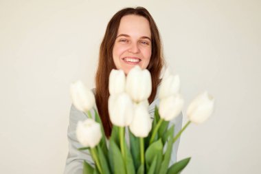 Bir kadın elinde bir buket beyaz lale tutarak neşeli bir gülümseme sergiliyor. Ayarlama, onun neşeli tavırlarını artıran tarafsız bir arkaplan içeriyor..