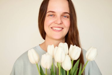 Bir kadın elinde taze bir buket beyaz lale tutarken neşeli bir ifadeyle ayakta duruyor. Arka plan hafifçe aydınlatılmış, neşeli tavrını geliştiriyor..