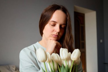 Bir kadın derin düşüncelere dalmış, bir buket beyaz laleye bakarken çenesini elinin üzerinde dinlendiriyor. Yumuşak ışık, sakin ve içe dönük bir atmosfer yaratır..