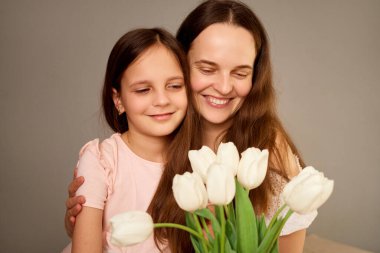 Bir anne ve küçük kızı, bir buket beyaz lalenin tadını çıkarırlarken, sıcak bir şekilde gülümsüyor. Onların sevgi dolu bağları bu iç açıcı anda açıkça görülüyor..