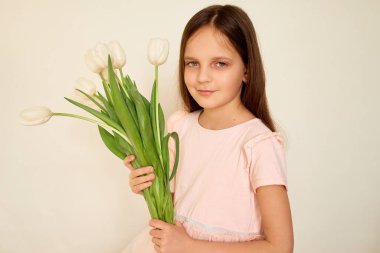 Elinde bir buket beyaz lale tutan genç bir kız hafifçe gülümsüyor. Açık pembe bir kıyafet giyiyor, yumuşak, nötr bir arka plana karşı duruyor, sakin bir atmosfer yaratıyor..