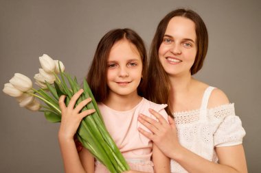 Bir anne ve kızı birlikte gülümsüyor, kızı da elinde bir buket taze beyaz lale tutuyor. Mutluluk ve birliktelik duygusu yayıyorlar..