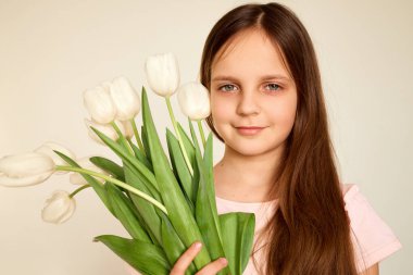 Genç bir kız bir buket taze beyaz lale tutarken nazikçe gülümsüyor. Açık renkli bir ortamda duruyor, sakinlik ve neşe saçıyor..