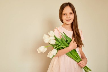 Bir kız, narin pembe bir elbise giymiş ve elinde bir buket taze beyaz lale tutarak aydınlık bir odada duruyor. Neşeli ifadesi mutluluğu ve masumiyeti yansıtıyor..