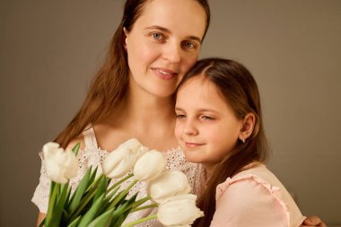 Bir kadın ve küçük kızı, beyaz lale buketi sergileyerek birbirlerine sımsıkı sarılıyorlar. İfadeleri, sevgi ve sevgiyi, tarafsız ışıklandırmayla kapalı bir ortamda, samimi bir ortamda iletir..