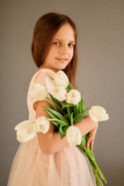 Açık pembe elbiseli genç bir kız elinde bir buket taze beyaz lale ile kendinden emin duruyor. Tarafsız arka plan onun zarif duruşunu ve neşeli ifadesini güçlendirir..