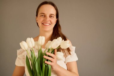 Bir kadın elinde canlı bir buket beyaz lale tutarak evde duruyor. Doğal ışık odayı dolduruyor, sıcak gülümsemesini ve çiçeklerin tazeliğini arttırıyor..