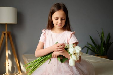 Yumuşak pembe elbiseli genç bir kız, şefkatle beyaz lale buketi tutuyor. Yeşillik ve doğal ışıkla dolu, parlak, modern bir oturma odasında duruyor..