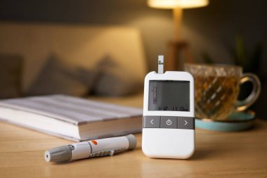 Bir glukometre kan şekeri seviyesini gösterirken test için bir kalem yakınlarda duruyor. Bir fincan çay ve açık bir kitap iyi ışıklandırılmış odanın huzurlu atmosferine ilave eder..