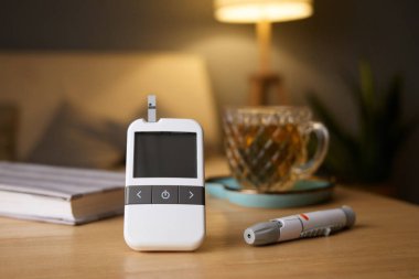 A glucometer with a test strip and a pen for insulin is placed on a wooden table in a cozy living room. A cup of tea and a notebook are in the background, creating a warm atmosphere.