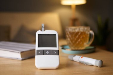 Tahta bir masanın üzerine kan şekeri testi sonuçlarını gösteren bir glukometre yerleştiriliyor. Yakınlarda, bir fincan çay ve veri toplamak için bir kalem rahat bir yaşam alanında rahatlatıcı bir atmosfer yaratır..