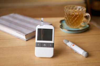 Bir glukometre bir defter ve bir fincan çayın yanındaki ahşap masada oturur. Yakın bir yerde bulunan bir kalem, evdeki kişisel sağlık durumunu gösteriyor..