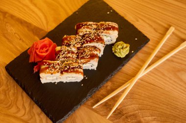 Sushi dürümleri, zencefil ve wasabi eşliğinde siyah bir tabakta ustalıkla düzenlenir. Sıcak ahşap masa öğle veya akşam yemeği sırasında rahatlatıcı bir atmosfer yaratır..