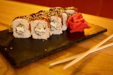 Lezzetli suşi ruloları, akşam yemeği sırasında modern bir restoranda, turşulu zencefil ve çubuklarla birlikte siyah bir tabakta güzelce düzenlenir..