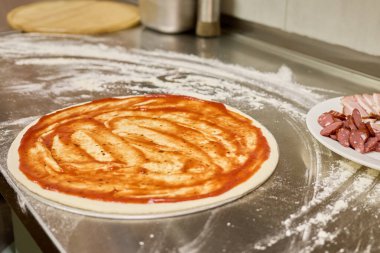 Taze pizza hamuru paslanmaz çelik bir yüzeye yayılır, domates sosuyla kaplanır. Yakınlarda, et dilimleri eklenmeyi bekliyor. Yoğun bir mutfak atmosferi sergiliyorlar..