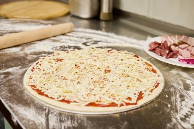 Hamur un tozlu bir tezgahın üzerine serpilmiş, üstüne domates sosu ve dilimlenmiş peynir serpiştirilmiş, dolgun bir mutfakta ilave malzemeler için hazır..