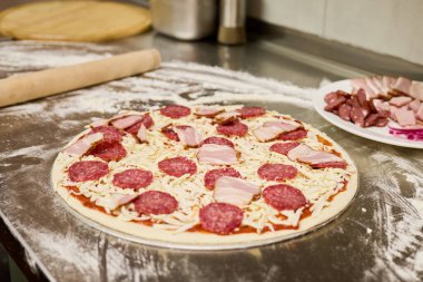 Sıcak bir mutfakta, ev yapımı pizza hamuru domates sosu, peynir, salam ve jambonla kaplanır, pişirilmeye hazırdır. Taze malzemeler, mutfak deneyimini geliştirmek için yakınlarda sergileniyor..