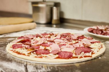 Taze yapılmış pizza hamuru un kaplı bir tezgaha serilir, üzerine et ve peynir karışımı konur, sıcak bir mutfakta pişirilmeye hazırdır..