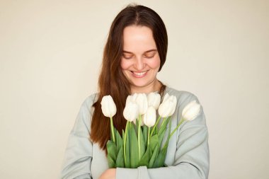 Neşeli bir kadın elinde bir buket beyaz laleyle mutluluk saçan bir ifadeyle ayakta duruyor. Arka plandaki hafif ton, sakin atmosferi güçlendirir..
