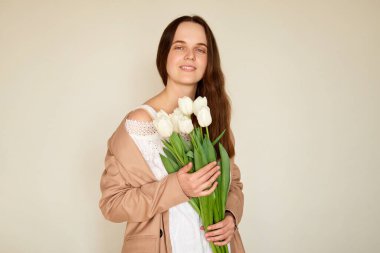 Genç bir kadın, yumuşak, aydınlık bir alanda, beyaz lalelerle dolu bir buketi sallayarak zarif bir şekilde duruyor. Rahat bir kıyafet giymiş, sakin bir atmosfer yayıyor..