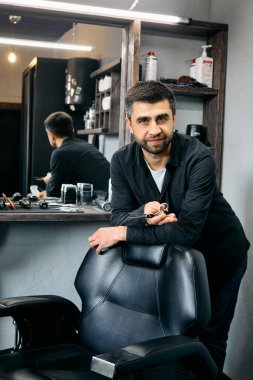 Yetenekli bir kuaför şık bir berber dükkanında berber koltuğunun yanında güvenle durur. Atmosfer sıcak, aletler ve ekipmanlarla dolu müşterilerin görünüşünü değiştirecek bir güne hazır..