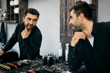 Bir kuaför aynadaki yansımasına bir sonraki saç kesimini düşünerek bakar. Şık berber dükkanında tezgahın üzerinde düzgün düzenlenmiş çeşitli alet ve ürünler var..