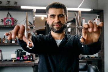 Bir kuaför, elinde makasla modern bir berber dükkanında güvenle duruyor ve saç kesimine hazırlanıyor. Şık iç mekan ve odaklanmış ifade müşteriler için davetkar bir ortam yaratır..