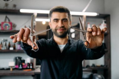 Yetenekli bir kuaför, canlı bir berber dükkanında şık saç kesimleri yapmaya hazır iki çift makas tutar. Mağazada moda bir atmosfer ve çeşitli saç ürünleri sergileniyor.