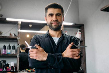 Kuaför şık bir berber dükkanında iki elinde de makasıyla güvenle durur. Parlak ışıklandırma, saç bakım ürünleriyle dolu iyi örgütlenmiş çalışma alanını aydınlatıyor.