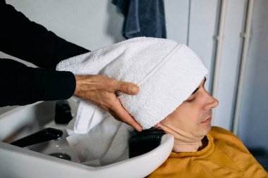 Çağdaş bir berber dükkanında, bir stilist müşterisinin saçını lavaboda dikkatlice yıkar, saç kesimine hazırlanır. Atmosfer rahat ve bakımlı..