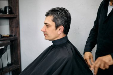 Kuaför saçını şekillendirirken berber koltuğunda pelerin giyen bir adam oturuyor. Atmosfer rahat, aletler düzgünce dizilmiş, profesyonel bir ortam yaratıyor..
