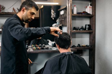 Yetenekli bir kuaför çağdaş bir berber dükkanında müşterinin saçını dikkatlice keser. Atmosfer canlı, araçlar ve ürünler yakınlarda düzenli bir şekilde düzenlenmiş, profesyonel bir ortam sergiliyor..