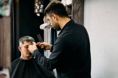 Yetenekli bir kuaför, çağdaş bir berber dükkanında müşterisinin saçını keserken dikkatle odaklanır. Atmosfer rahatlatıcıdır. Dekorasyon ve aletler görünür, karşılama alanı oluşturur..