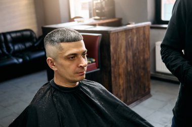 Bir kuaför, çağdaş bir berber dükkanında oturan odaklanmış bir müşteriye titiz bir saç kesimi yapar. Sıcak ortam tımar deneyimini geliştirir, profesyonel hizmeti ön plana çıkarır..