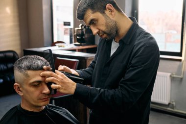 Profesyonel bir kuaför, iyi ışıklandırılmış bir yerdeki çağdaş bir berber dükkanında müşterinin saçını dikkatlice şekillendirir.. 