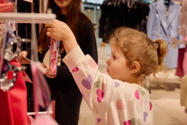 Kalp desenli süveter giyen genç bir kız, yaşlı bir refakatçi kalabalık bir alışveriş merkezinde bakarken askıdaki kıyafetleri inceliyor. Atmosfer canlı ve renkli..