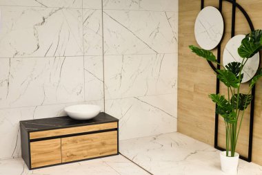 Şık bir banyoda zarif mermer fayanslar ön çekmeceleri olan minimalist ahşap kibir ve yemyeşil bir bitkiyi yansıtan dairesel bir ayna sergilenir..