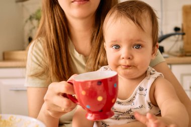 Bir bebek mutfak masasında otururken renkli bir fincan tutar. Arka planda bir bakıcı beliriyor. Atıştırma sırasında sıcak bir aile ortamı yaratıyor..