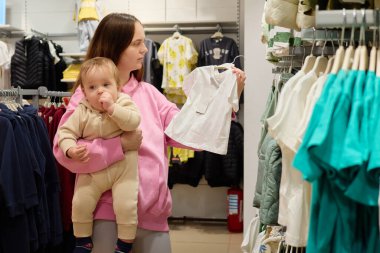 Bir anne, bir perakende mağazasında bebek kıyafetlerini karıştırırken bebeğini yumuşak bir taşıyıcıda taşır. Seçimini göz önünde bulundurarak beyaz bir elbiseyi inceliyor..
